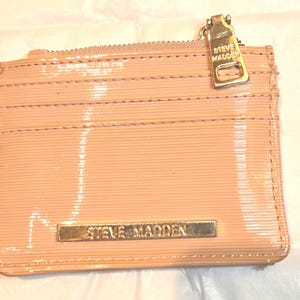 May include: A blush-colored Steve Madden cardholder with a glossy finish. The rectangular wallet features a zipper closure, multiple card slots, and a gold-tone metal zipper pull and logo. The Steve Madden logo is displayed on a gold-tone metal plate.