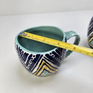 May include: A ceramic mug with a teal interior and handle, and a white, navy, and yellow geometric pattern on the exterior. The mug is approximately 4 inches wide.