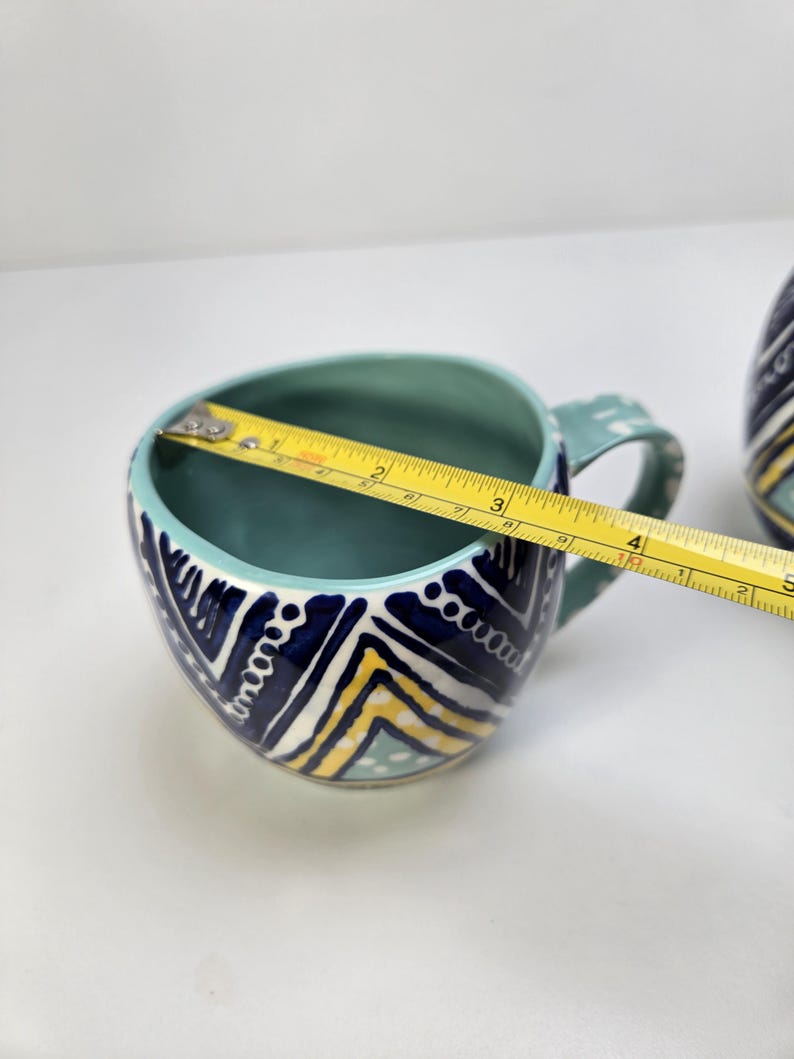 May include: A ceramic mug with a teal interior and handle, and a white, navy, and yellow geometric pattern on the exterior. The mug is approximately 4 inches wide.
