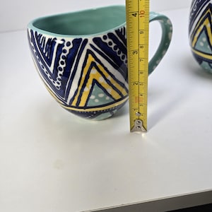 May include: A ceramic mug with a teal interior and handle. The exterior features a geometric pattern in navy blue, yellow, and white. The mug is approximately 4 inches tall, as measured by a yellow tape measure.