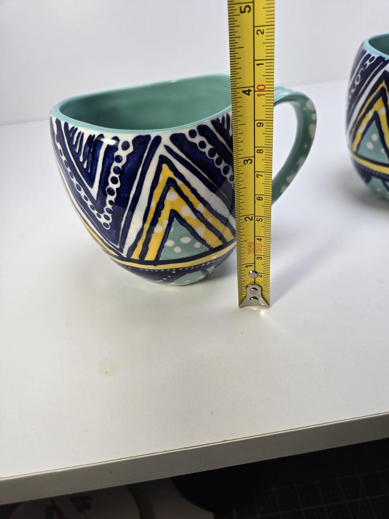 May include: A ceramic mug with a teal interior and handle. The exterior features a geometric pattern in navy blue, yellow, and white. The mug is approximately 4 inches tall, as measured by a yellow tape measure.
