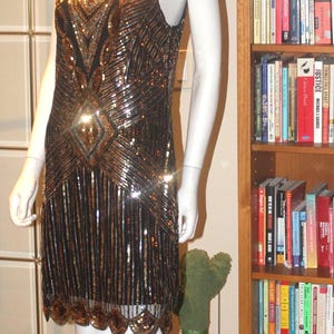 May include: A sleeveless black dress featuring intricate gold and bronze sequin patterns in a geometric design. The dress has a scoop neckline and a scalloped hemline with dangling fringe-like sequin details, creating a shimmering effect.