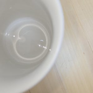 May include: A white ceramic mug with a smooth, glossy interior. The mug is set against a light-colored wooden surface. The interior of the mug reflects light, creating highlights.