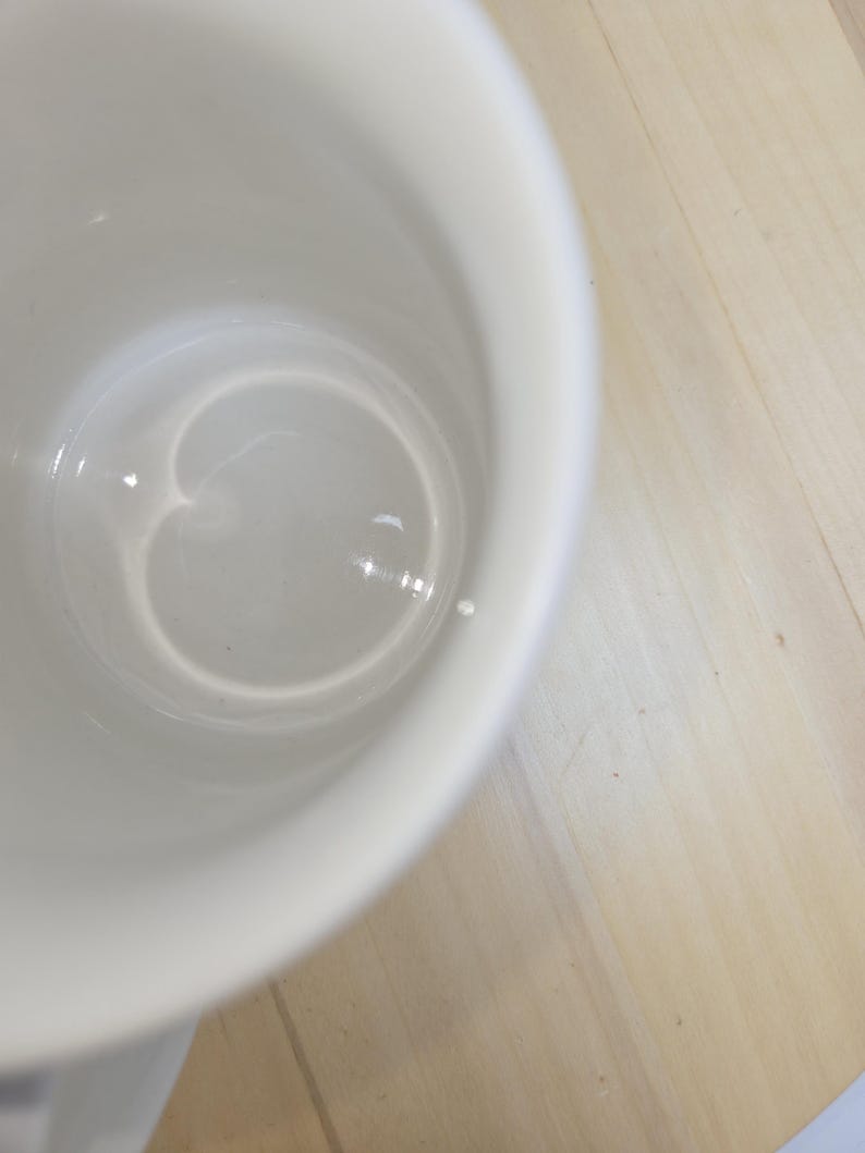 May include: A white ceramic mug with a smooth, glossy interior. The mug is set against a light-colored wooden surface. The interior of the mug reflects light, creating highlights.