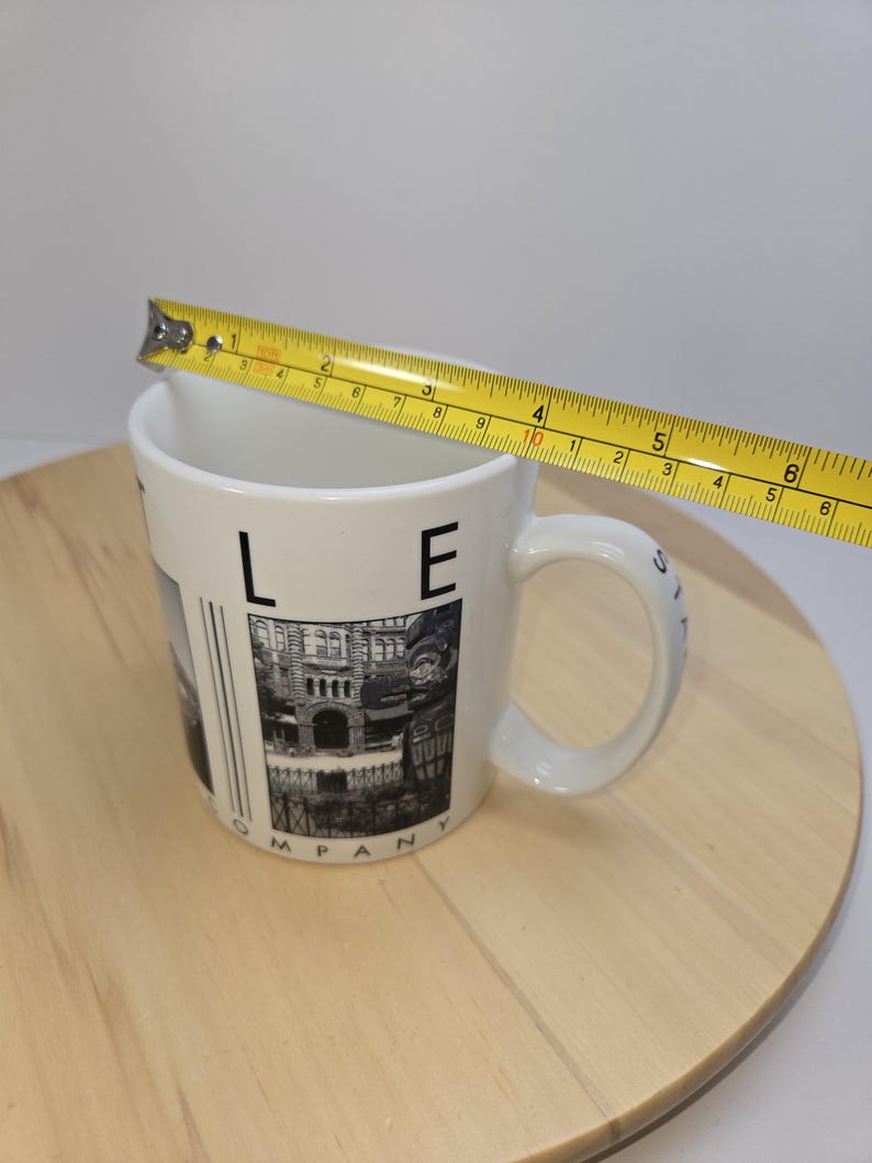 May include: White ceramic mug with a black and white image of a building and the text "LE COMPANY". The mug is approximately 4 inches in diameter and has a white handle.