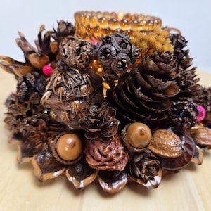 May include: A decorative candle holder made from natural elements. The base is composed of pine cones, acorns, and nuts, with a small amber glass candle cup in the center. Pink beads add a touch of color.