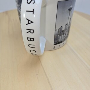 May include: Cream-colored ceramic mug with the word "STARBUCKS" printed in black on the handle. The mug features a black and white image of a city skyline with a tall tower. The mug is on a light wood surface.