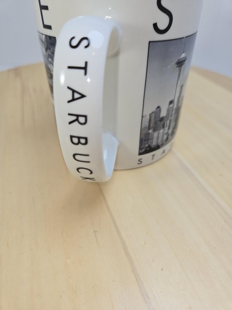 May include: Cream-colored ceramic mug with the word "STARBUCKS" printed in black on the handle. The mug features a black and white image of a city skyline with a tall tower. The mug is on a light wood surface.