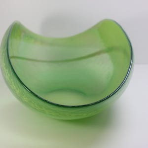 Può includere: Una ciotola in vetro verde chiaro traslucido con un bordo curvo e asimmetrico. La ciotola presenta una superficie liscia e lucida e una sottile trama interna che crea un aspetto morbido e stratificato all'interno della struttura in vetro.