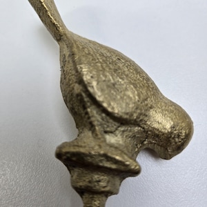 May include: A brass-colored bird figurine with a detailed, textured surface. The bird is perched atop a decorative base, and the tail is angled upwards. The figurine is likely a decorative item or part of a larger piece.