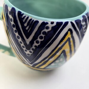 May include: A ceramic mug with a teal interior and handle. The exterior features a navy blue and white chevron pattern with yellow accents. The mug has a rounded shape and a wide opening, suitable for coffee or tea.