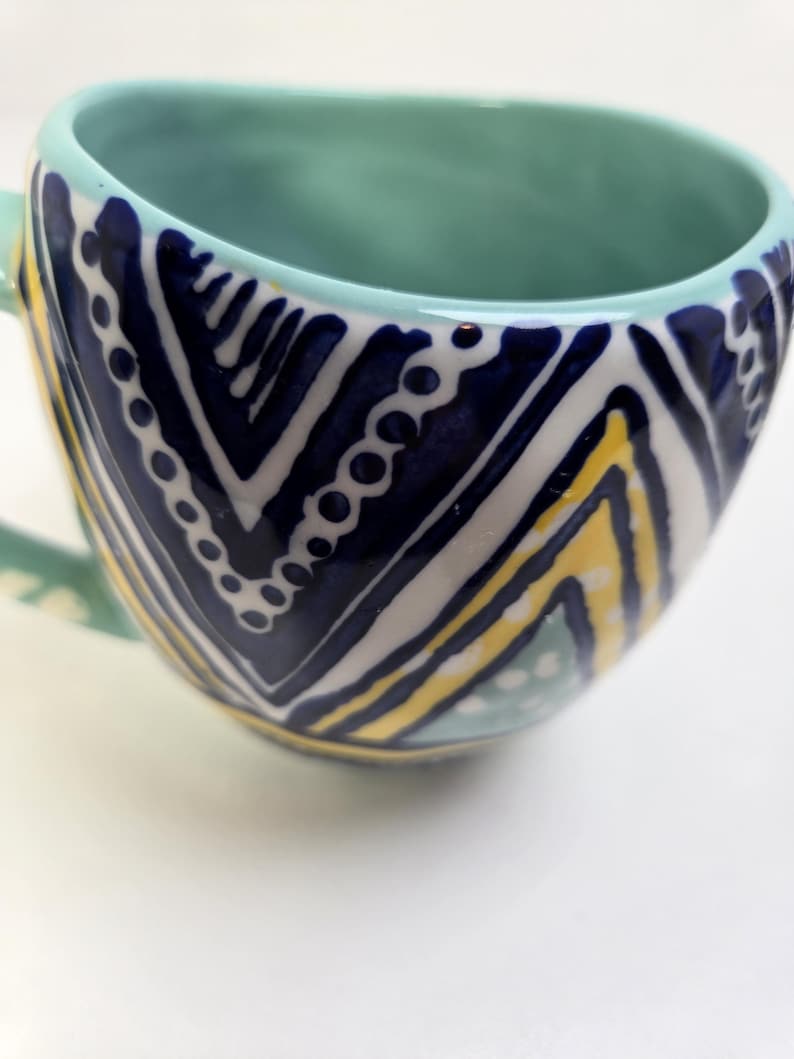 May include: A ceramic mug with a teal interior and handle. The exterior features a navy blue and white chevron pattern with yellow accents. The mug has a rounded shape and a wide opening, suitable for coffee or tea.