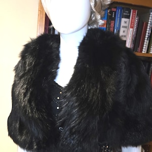 Puede incluir: Un chal de piel sintética negra con un cuello ancho, exhibido en un maniquí. El chal está superpuesto a un vestido negro con detalles de lentejuelas doradas. El maniquí tiene una peluca rubia.