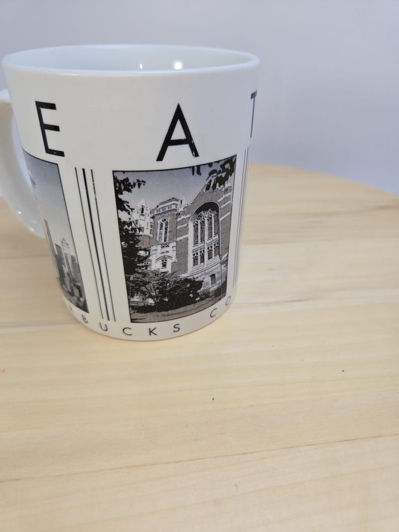 May include: White ceramic mug featuring a black and white image of a building and the word "EAT" on the side. The mug has a handle and the words "BUCKS CO" are printed at the bottom. The mug is sitting on a light brown wooden surface.