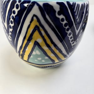 May include: A ceramic bowl with a turquoise interior and a navy, white, and yellow geometric pattern on the exterior. The bowl has a unique shape and a glossy finish. The design includes chevron and dotted patterns.