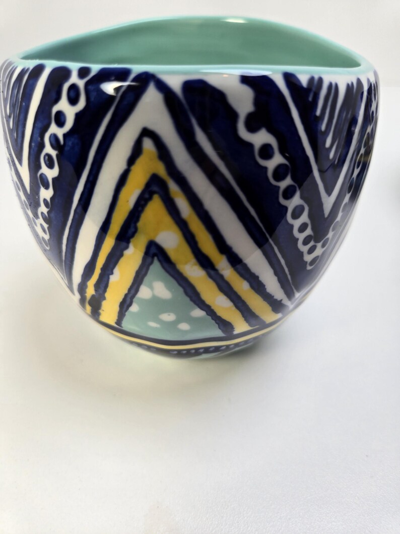May include: A ceramic bowl with a turquoise interior and a navy, white, and yellow geometric pattern on the exterior. The bowl has a unique shape and a glossy finish. The design includes chevron and dotted patterns.