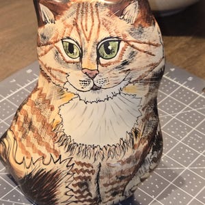 May include: A hand-painted ceramic bottle shaped like a seated cat. The cat has green eyes, a pink nose, and brown and tan fur with detailed stripes. The bottle has a cream-colored neck and a dark stopper.
