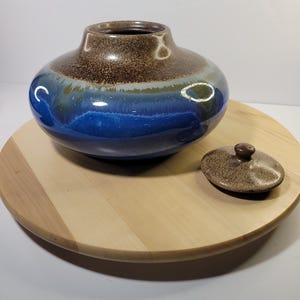 Op de afbeelding: Een blauwe en bruine keramische pot met een deksel. De pot heeft een brede opening en een rond lichaam. Het deksel is bruin met een knop bovenop.