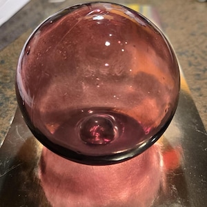 May include: A spherical glass float in a deep translucent purple hue. The surface of the orb is smooth and clear, featuring subtle internal air bubbles and a distinct circular indentation at the base, reflecting light across its rounded form.