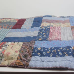 May include: A rectangular patchwork quilt constructed from various fabric strips in shades of blue, pink, and burgundy. The textile features a mix of floral, paisley, and solid patterns stitched together with visible quilting lines throughout the surface.