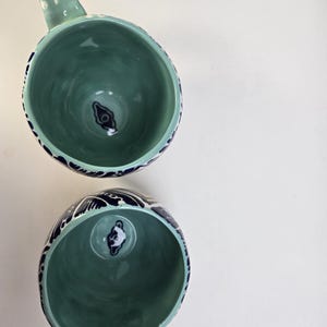 May include: Two ceramic mugs with a teal interior and a teal handle. The exterior rim is decorated with a dark blue wave pattern. The bottom of the mug has a dark blue and white design. The mugs are on a white background.