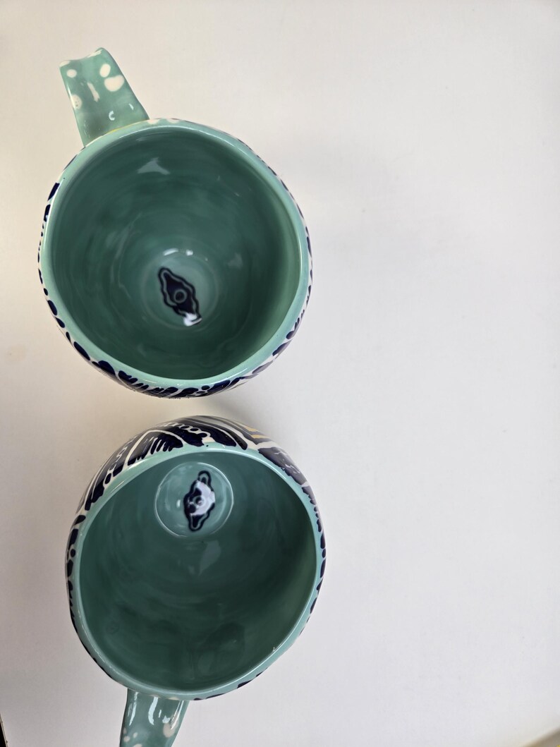 May include: Two ceramic mugs with a teal interior and a teal handle. The exterior rim is decorated with a dark blue wave pattern. The bottom of the mug has a dark blue and white design. The mugs are on a white background.