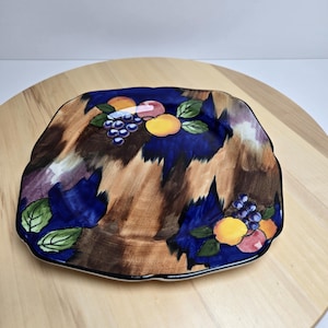 Puede incluir: Plato cuadrado de cerámica con diseño de frutas. El plato presenta un fondo marrón y azul con ilustraciones de uvas, melocotones y hojas verdes. El plato tiene un borde negro y está sobre una superficie de madera.