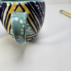 May include: A ceramic mug with a teal interior and handle. The exterior features a geometric pattern in navy blue, yellow, and white. The handle is decorated with white polka dots. The mug is designed with a unique shape.