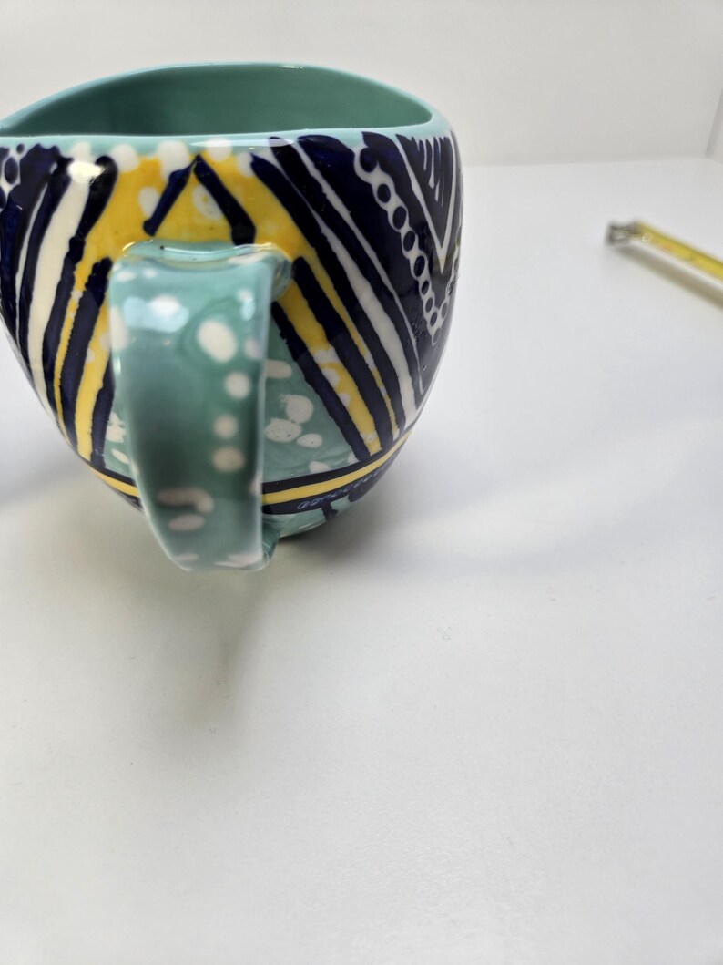 May include: A ceramic mug with a teal interior and handle. The exterior features a geometric pattern in navy blue, yellow, and white. The handle is decorated with white polka dots. The mug is designed with a unique shape.