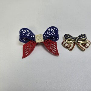 May include: Two patriotic bow-shaped brooches. One is a blue, red, and white openwork design. The other is a smaller bow with a gold center, and red, white, and blue stripes and stars.
