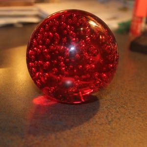 May include: A spherical glass paperweight in a deep translucent red color. The interior of the sphere is filled with numerous small, suspended air bubbles of varying sizes, creating a textured, effervescent appearance throughout the solid glass.