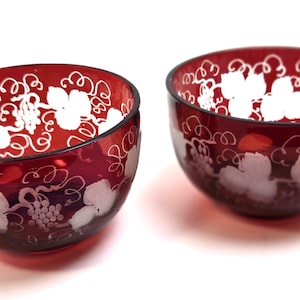 May include: Two deep red glass bowls featuring a white etched design of grape clusters and swirling vine patterns. The bowls have a rounded shape and a smooth, translucent finish, showcasing the contrast between the dark red glass and white details.