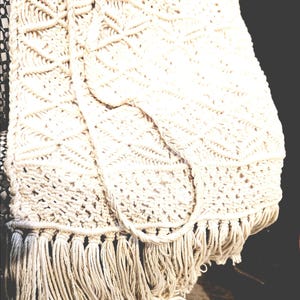 May include: Cream-colored macrame bag with a geometric pattern and long fringe. The bag has a woven design and a long strap. The bag is made of a soft, natural fiber.