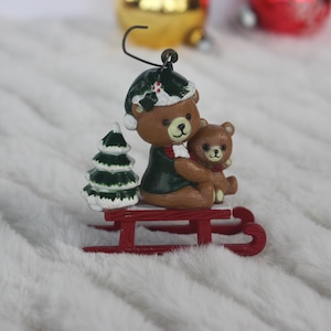 May include: A Christmas ornament featuring two brown teddy bears on a red sled. The larger bear is wearing a green and white hat and holding a smaller bear. A green pine tree is in the background.
