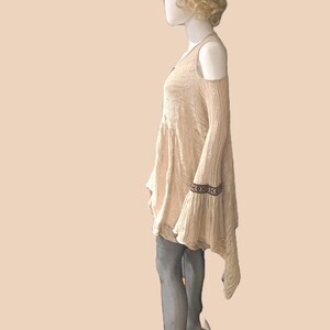Op de afbeelding: Beige jurk met open schouders, lange mouwen en een uitlopende rok. De jurk heeft een decoratieve band op de mouwboorden. De mannequin draagt zwarte visnetkousen.