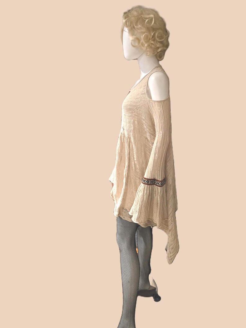 Op de afbeelding: Beige jurk met open schouders, lange mouwen en een uitlopende rok. De jurk heeft een decoratieve band op de mouwboorden. De mannequin draagt zwarte visnetkousen.