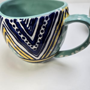 May include: A ceramic mug with a teal interior and handle. The exterior features a geometric pattern in navy blue, white, and yellow. The handle is decorated with white polka dots. This handmade mug is perfect for coffee or tea.
