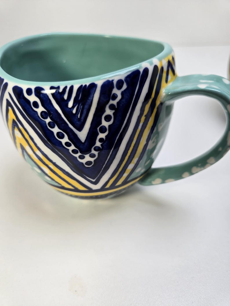 May include: A ceramic mug with a teal interior and handle. The exterior features a geometric pattern in navy blue, white, and yellow. The handle is decorated with white polka dots. This handmade mug is perfect for coffee or tea.