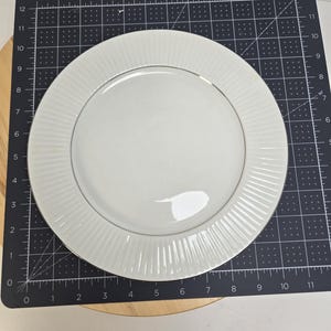 May include: A white ceramic plate with a silver rim and a textured, ridged design on the outer edge. The plate is centered on a black cutting mat with inch measurements.