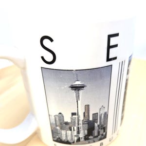 May include: White ceramic mug featuring a black and white image of the Seattle skyline, including the Space Needle. The mug has the letters "S" and "E" printed on the side, with the word "STARBUCKS" partially visible.