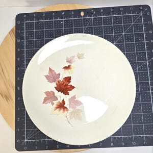 May include: Cream-colored ceramic plate with a decorative autumn leaf design in shades of red, pink, and beige. The plate is round and appears to be approximately 10 inches in diameter. The leaves are clustered on the left side of the plate.