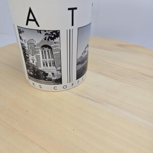 May include: White ceramic coffee mug with black text and images. The mug features the letters "A T" and the words "KS COFFEE" in a circular pattern. The mug has two black and white images, one of a building and the other of a mountain.