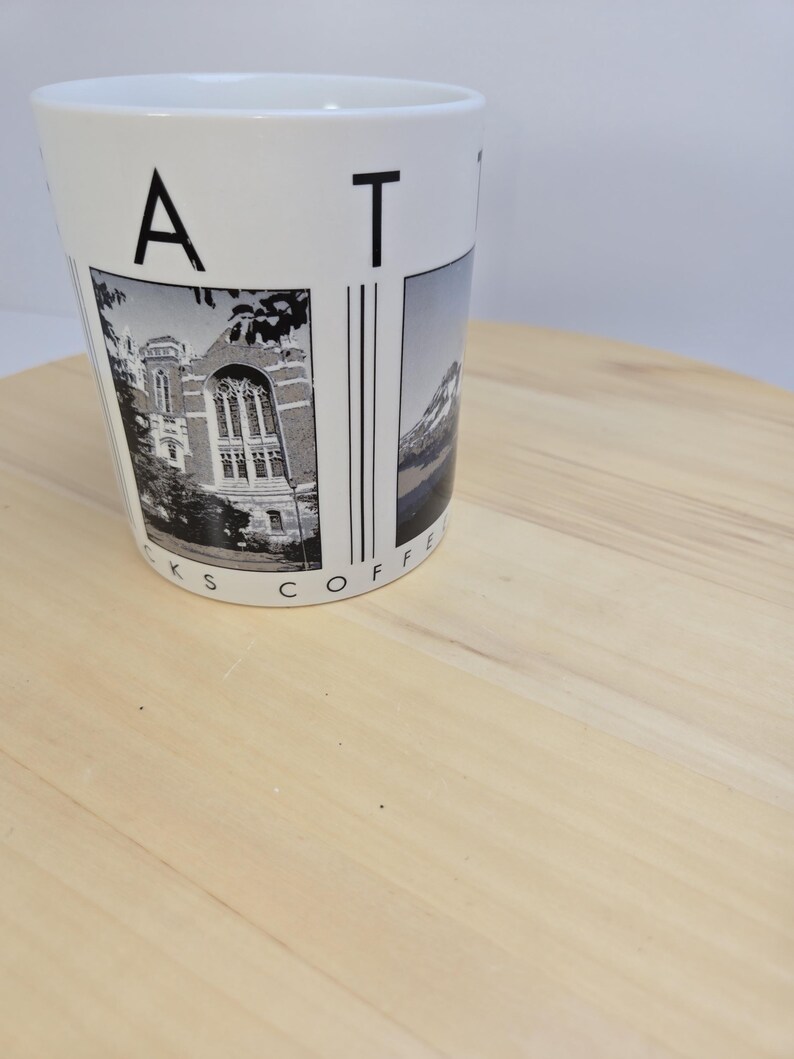 May include: White ceramic coffee mug with black text and images. The mug features the letters "A T" and the words "KS COFFEE" in a circular pattern. The mug has two black and white images, one of a building and the other of a mountain.