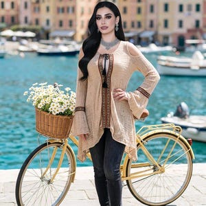 May include: A beige open-shoulder tunic with bell sleeves, featuring a decorative trim and a tassel tie. The tunic is paired with dark jeans. A yellow bicycle with a wicker basket filled with white flowers is in the foreground.