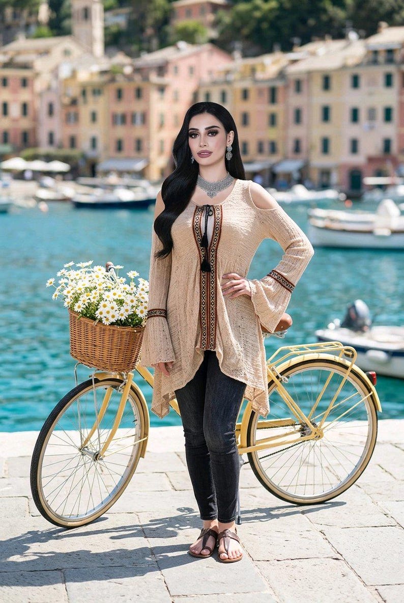 Op de afbeelding: Een beige tuniek met open schouders en klokmouwen, met een decoratieve bies en een kwastje. De tuniek wordt gecombineerd met een donkere jeans. Een gele fiets met een rieten mand vol witte bloemen staat op de voorgrond.