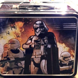May include: A black metal lunch box featuring a graphic of three Star Wars stormtroopers in white armor with blasters. The background shows an orange and black explosion scene with TIE fighters. The word THERMOS is printed in the bottom left corner.