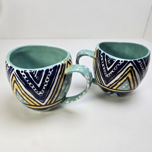 May include: Two ceramic mugs with teal interiors and handles. The exterior features a navy blue, yellow, and white geometric pattern. The mugs are on a white surface.