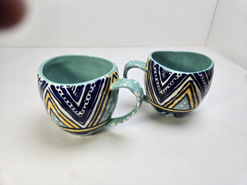 May include: Two ceramic mugs with teal interiors and handles. The exterior features a navy blue, yellow, and white geometric pattern. The mugs are on a white surface.