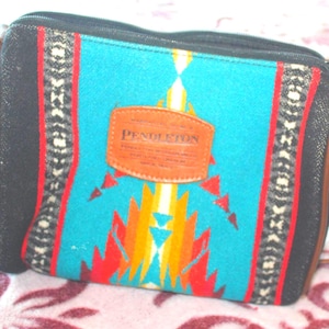 May include: A rectangular crossbody bag with a turquoise, red, and yellow geometric pattern. The bag has black sides and a brown leather strap. A brown leather patch with the brand name "Pendleton" is centered on the front.