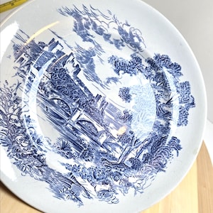 Puede incluir: Plato de cerámica blanca con un diseño de paisaje azul y blanco. El plato presenta una ilustración detallada de un puente, árboles y edificios. El plato tiene un aspecto ligeramente desgastado.